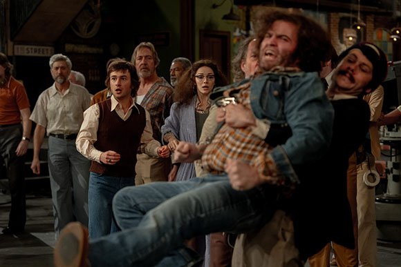 A group of people looking on with shocked and worried expressions in a dimly lit indoor setting.