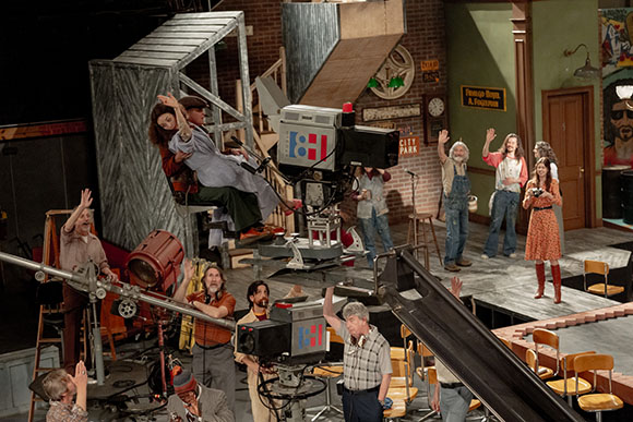 Actors and crew members pose on a theatrical stage set designed to look like a film production.