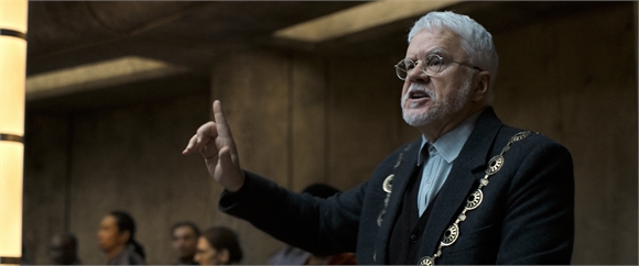 An older man with white hair and glasses gestures with one finger raised while speaking.