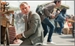 A man in a grey suit sits on the ground near a street vendor's stall in a busy outdoor area.