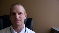A man with short hair and a white polo shirt sits in a black chair against a brown wall.