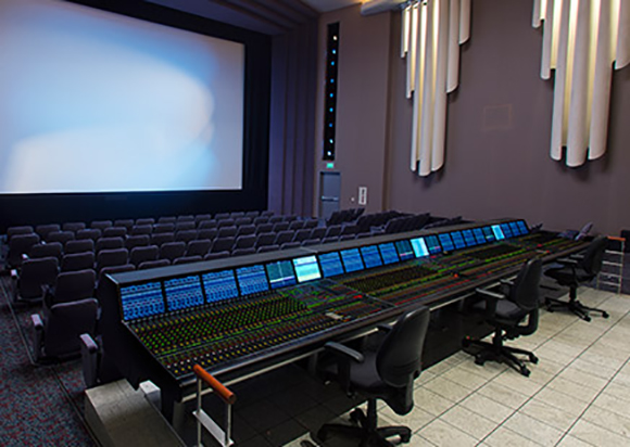 A long, complex control panel with many buttons and screens sits before a large cinema screen.