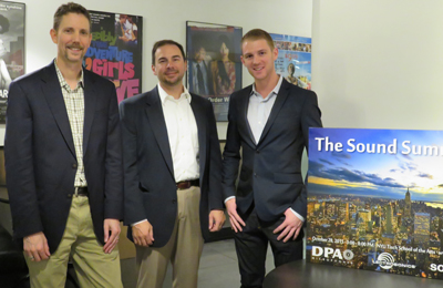 Three men in business casual attire standing in an office next to a large blue poster.