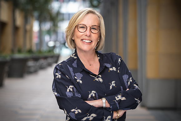 A smiling blonde woman wearing glasses and a dark patterned shirt stands with her arms crossed outdoors.