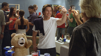 A man in a white t-shirt smiles and raises a shot glass during a crowded indoor party.