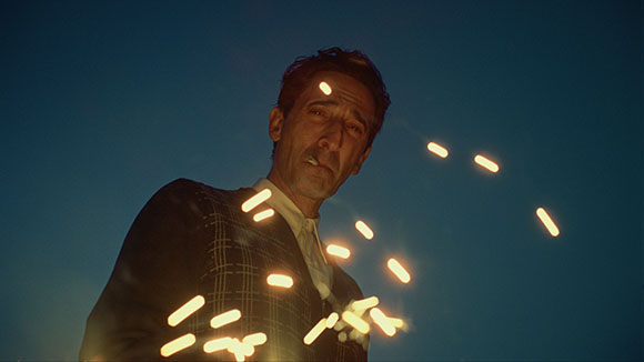 A man in a dark patterned suit looks upward against a dark blue sky as bright orange sparks fly past him.