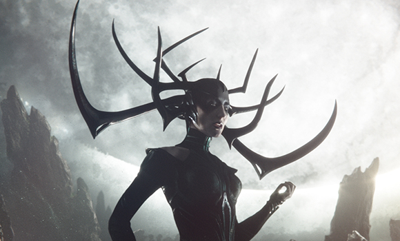 A woman wearing dark armor and a large, intricate black horned headpiece against a bright, cloudy sky.