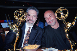 Two men smiling while seated next to two gold Emmy awards.