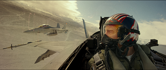A pilot in a cockpit wearing a helmet and oxygen mask, flying near another fighter jet over mountains.