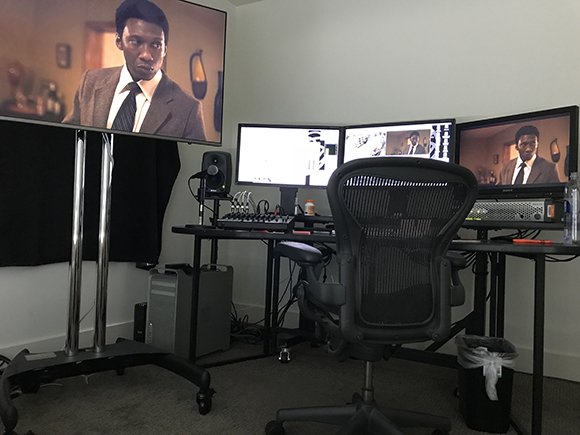 A video editing setup with three monitors, a black mesh office chair, and a computer tower.