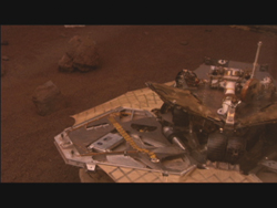 A robotic rover sits on a landing platform amidst a reddish, rocky landscape.