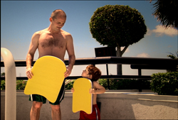 A shirtless man and a young child stand outdoors holding large yellow foam boards.