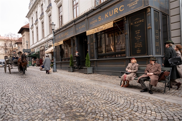 People in vintage clothing walk and sit on a cobblestone street past a dark storefront with gold lettering.