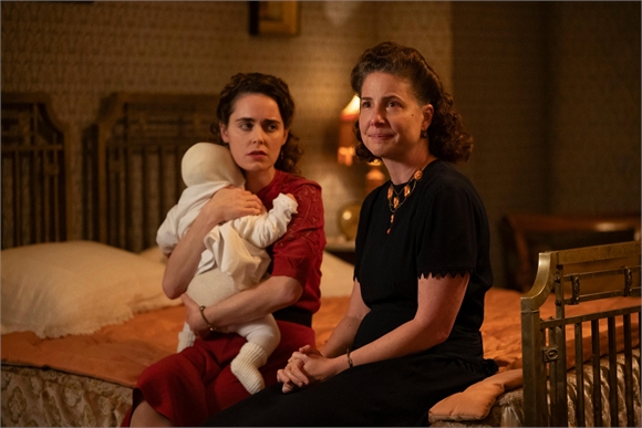 A woman in a red dress holds a white swaddled baby while another woman in black sits beside her on a bed.
