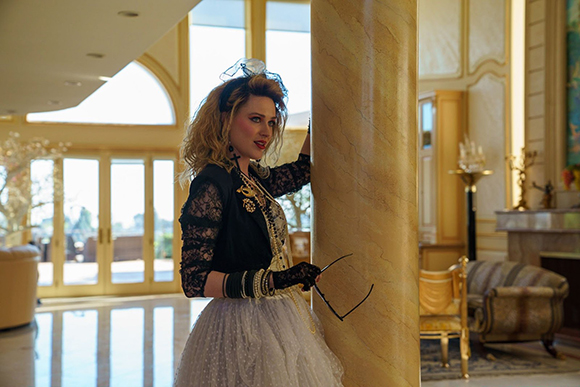 A woman with blonde hair wearing a black lace top and white tulle skirt leans against a large marble pillar.