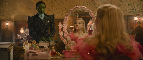 A green-skinned woman in a dark pinstripe suit stands behind a blonde woman wearing pink in a decorated room.