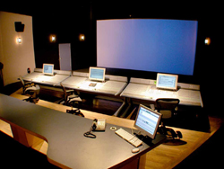 A dark control room featuring several computer monitors on desks facing a large blue projection screen.