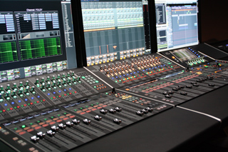 A large digital audio mixing console with numerous sliders, knobs, and glowing buttons in front of computer monitors.