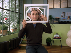 A man holds a white frame in front of his face, revealing several reflections of his own face within it.