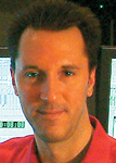A man with dark hair wearing a red shirt, looking forward against a dark background.