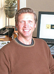 A smiling man with short brown hair wearing a brown sweater sits in front of a computer monitor.