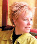 A woman with short, spiky blonde hair wearing a yellow jacket and looking off-camera.