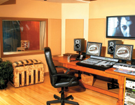 A recording studio control room with a large mixing console, monitors, a computer, and a view into a live room.
