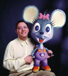 A man in a light yellow shirt smiles while sitting next to a large, cartoonish grey mouse puppet.