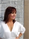 A woman with dark hair smiling while wearing a white V-neck blouse against a patterned background.