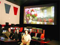 Three people sit at a desk in a dark control room facing a large screen displaying a floral image.
