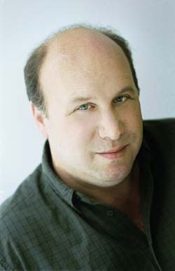 A man with thinning hair and blue eyes smiling slightly against a light background.